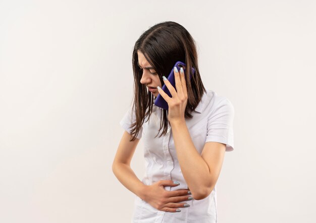 Chica joven con camisa blanca hablando por teléfono móvil que parece insalubre de pie sobre la pared blanca