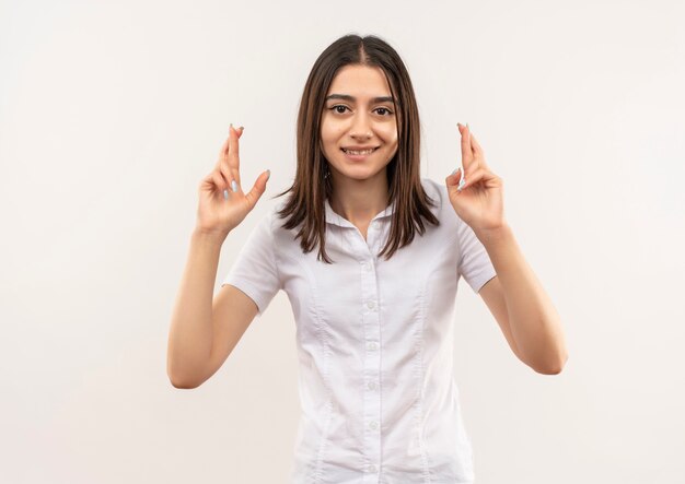 Chica joven con camisa blanca haciendo deseo deseable cruzar los dedos con expresión de esperanza de pie sobre la pared blanca