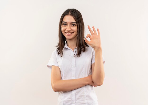 Chica joven con camisa blanca mirando al frente sonriendo mostrando signo ok de pie sobre la pared blanca