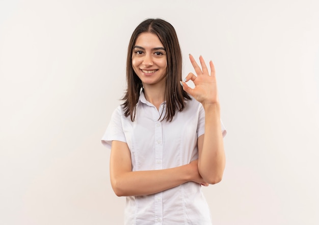 Chica joven con camisa blanca mirando al frente sonriendo mostrando signo ok de pie sobre la pared blanca