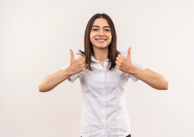 Chica joven con camisa blanca mirando hacia el frente sonriendo alegremente mostrando los pulgares para arriba de pie sobre la pared blanca