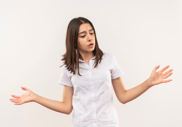 Chica joven con camisa blanca mirando incierto y confundido extendiendo los brazos a los lados de pie sobre la pared blanca