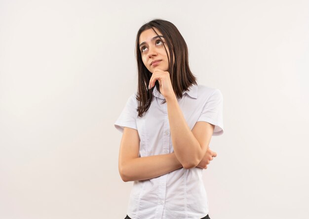 Chica joven con camisa blanca mirando a un lado con la mano en la barbilla perpleja de pie sobre la pared blanca