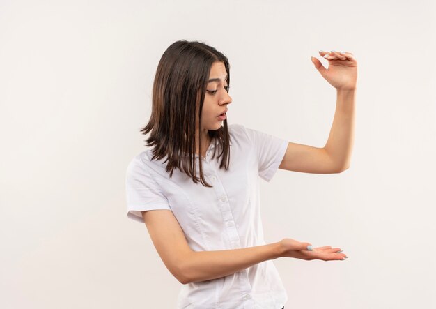 Chica joven con camisa blanca mirando a un lado mostrando gesto de tamaño con las manos, símbolo de medida de pie sobre la pared blanca