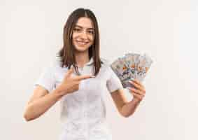 Foto gratuita chica joven con camisa blanca mostrando dinero en efectivo apuntando con el dedo a ella feliz sonriendo de pie sobre la pared blanca