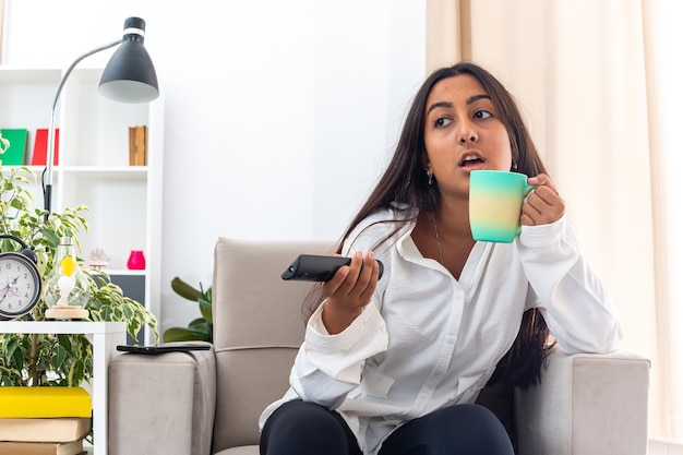 Chica joven con camisa blanca y pantalón negro con control remoto de tv sosteniendo la taza viendo la televisión sentado en la silla en la sala de luz