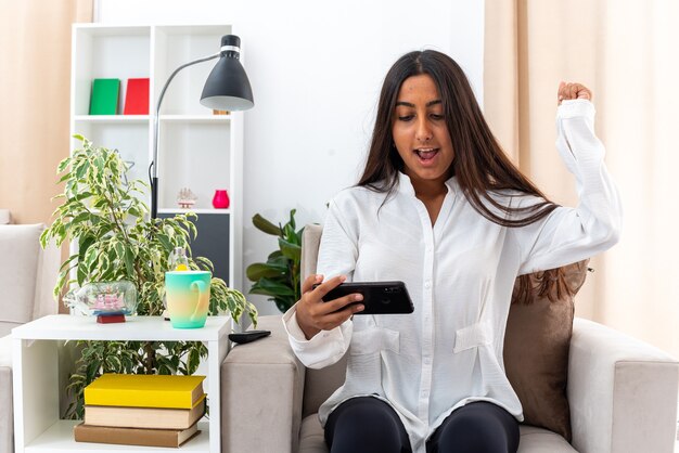 Chica joven con camisa blanca y pantalón negro jugando con su teléfono inteligente feliz y emocionada sentada en la silla en la sala de luz