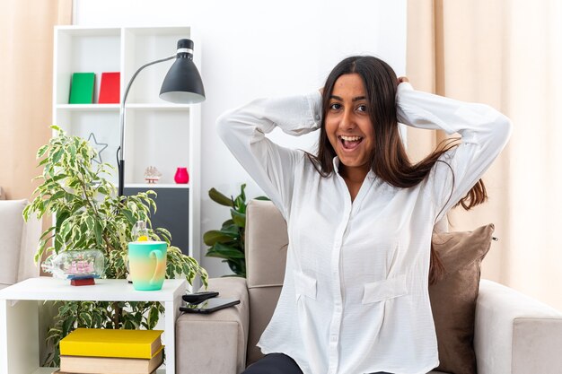 Chica joven con camisa blanca y pantalón negro mirando a la cámara feliz y emocionada tomados de la mano en la cabeza sentada en la silla en la sala de luz