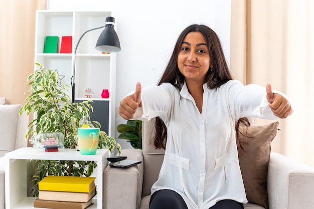 Chica joven con camisa blanca y pantalón negro mirando a la cámara feliz y positivo mostrando los pulgares para arriba sentado en la silla en la sala de luz