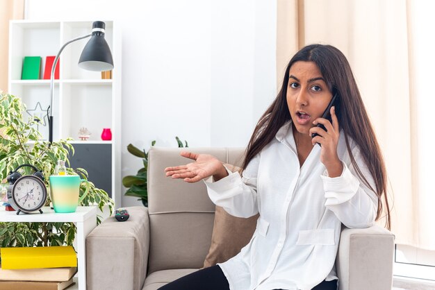 Chica joven con camisa blanca y pantalón negro que parece confundida y disgustada mientras habla por teléfono móvil sentado en la silla en la sala de estar luminosa