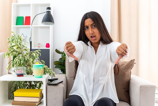 Chica joven con camisa blanca y pantalón negro con rostro serio disgustado mostrando los pulgares para arriba sentado en la silla en la sala de estar luminosa