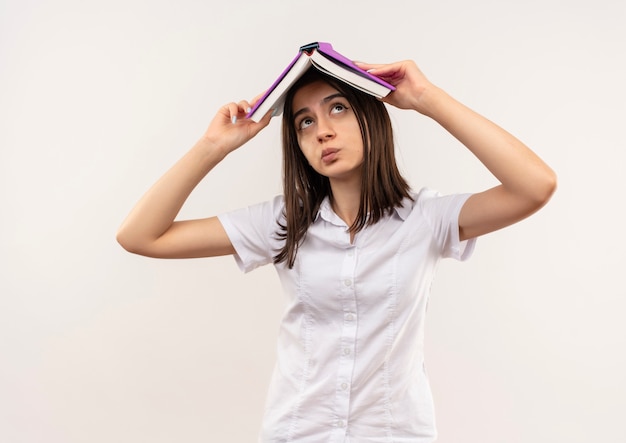 Chica joven con camisa blanca sosteniendo un libro abierto sobre su cabeza mirando confundido parado sobre la pared blanca