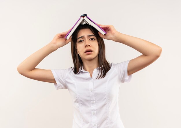 Chica joven con camisa blanca sosteniendo un libro abierto sobre su cabeza mirando confundido parado sobre la pared blanca