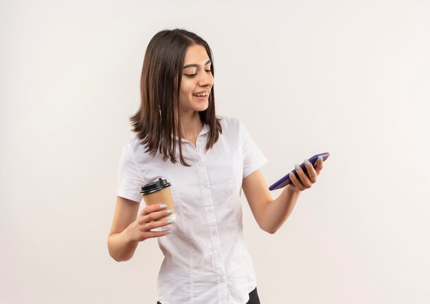 Chica joven en camisa blanca sosteniendo la taza de café mirando la pantalla de su móvil sonriendo alegremente de pie sobre la pared blanca