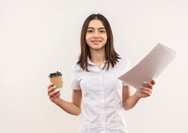 Chica joven con camisa blanca sosteniendo una taza de café y páginas en blanco sonriendo confiado de pie sobre la pared blanca