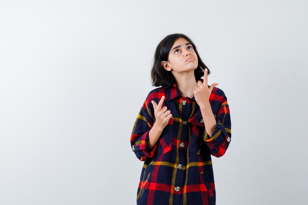 Chica joven en camisa a cuadros apuntando hacia arriba con los dedos índices y luciendo linda vista frontal.