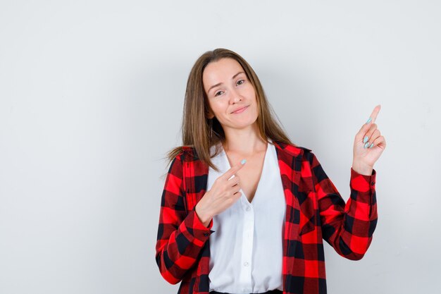 Chica joven en camisa a cuadros, blusa apuntando hacia arriba y mirando complacido, vista frontal.