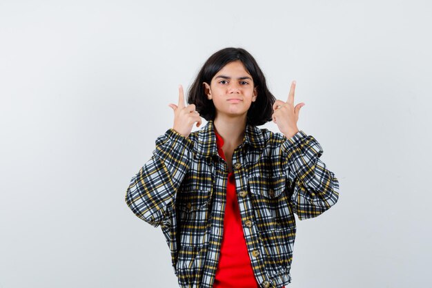 Chica joven en camisa a cuadros y camiseta roja apuntando hacia arriba y mirando linda vista frontal.