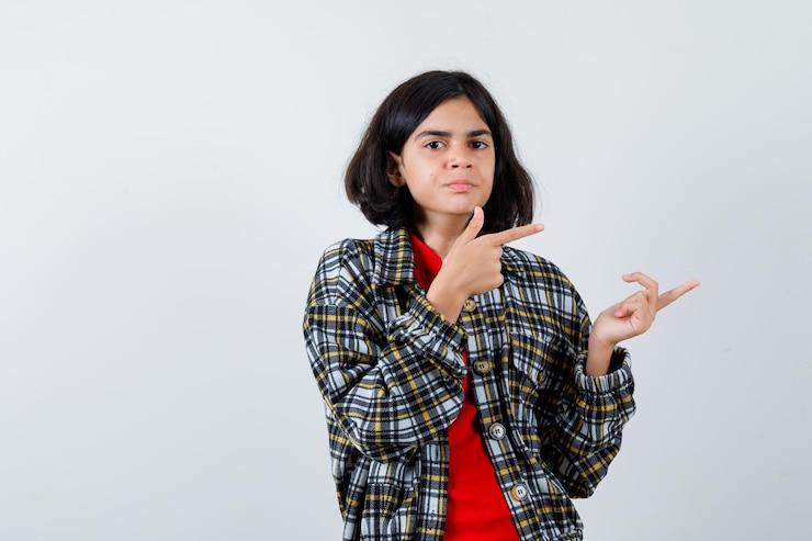 Foto gratuito chica joven con camisa a cuadros y camiseta roja apuntando a la derecha con los dedos índices y mirando seriamente, vista frontal.