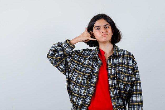 Chica joven en camisa a cuadros y camiseta roja que muestra el gesto de llamarme y mirando seria, vista frontal.