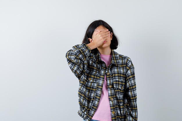 Chica joven en camisa a cuadros y camiseta rosa cubriéndose los ojos con la mano y mirando tímida, vista frontal.