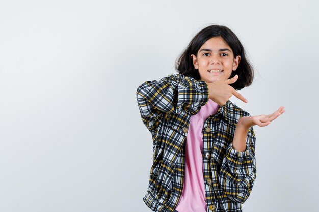 Chica joven en camisa a cuadros y camiseta rosa estirando una mano mientras sostiene algo y apuntando con el dedo índice y luciendo bonita, vista frontal.