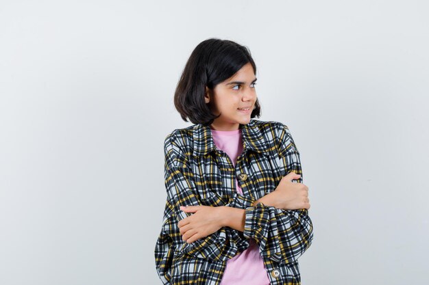 Chica joven en camisa a cuadros y camiseta rosa de pie con los brazos cruzados, mirando a otro lado y mirando enfocado, vista frontal.