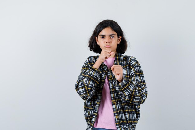 Chica joven en camisa a cuadros y camiseta rosa de pie en pose de boxeador y mirando serio, vista frontal.