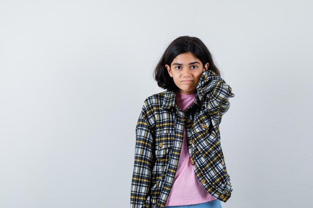 Chica joven en camisa a cuadros y camiseta rosa poniendo la mano detrás del cuello y luciendo bonita, vista frontal.