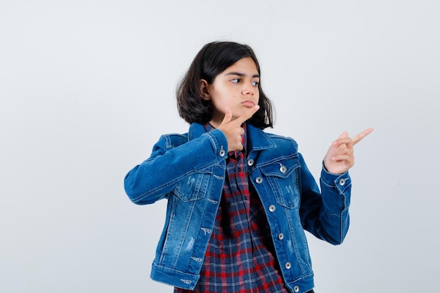 Chica joven en camisa a cuadros y chaqueta de jean apuntando hacia la derecha con los dedos índices y mirando seria