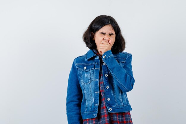 Chica joven en camisa a cuadros y chaqueta de jean cubriendo la boca con la mano, bostezando y con aspecto cansado