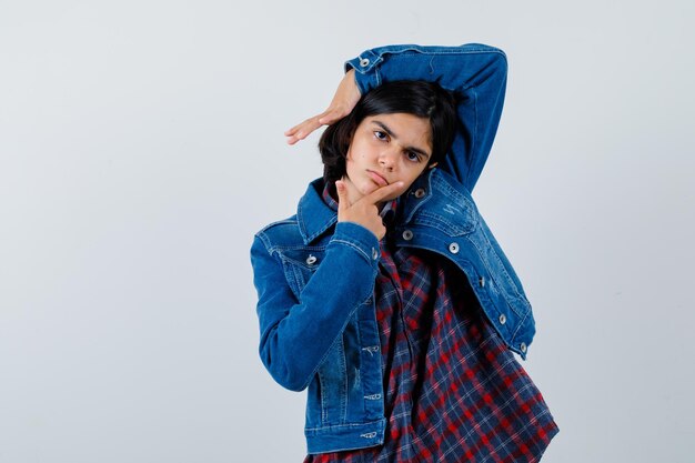 Chica joven en camisa de cuadros y chaqueta de jean poniendo el dedo índice en la barbilla mientras levanta la mano por encima de la cabeza y se ve bonita