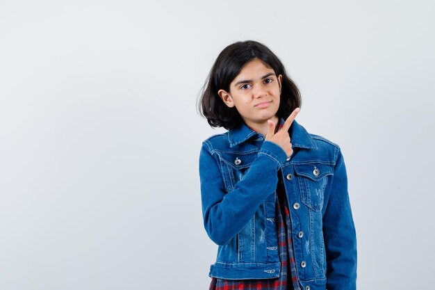 Chica joven con camisa a cuadros y chaqueta de mezclilla apuntando a la derecha con el dedo índice y mirando serio, vista frontal.