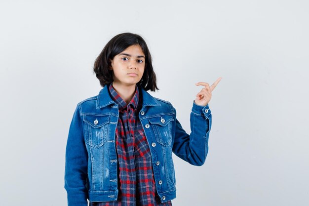 Chica joven en camisa a cuadros y chaqueta de mezclilla apuntando a la derecha y mirando seria, vista frontal.