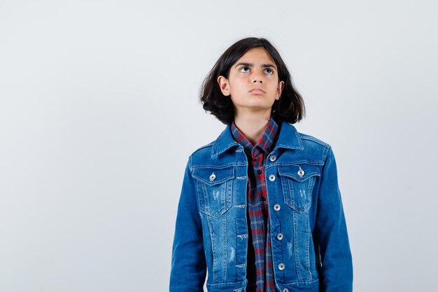 Foto gratuita chica joven en camisa a cuadros y chaqueta de mezclilla de pie con la espalda recta, mirando hacia arriba y posando a la cámara y mirando pensativo, vista frontal.