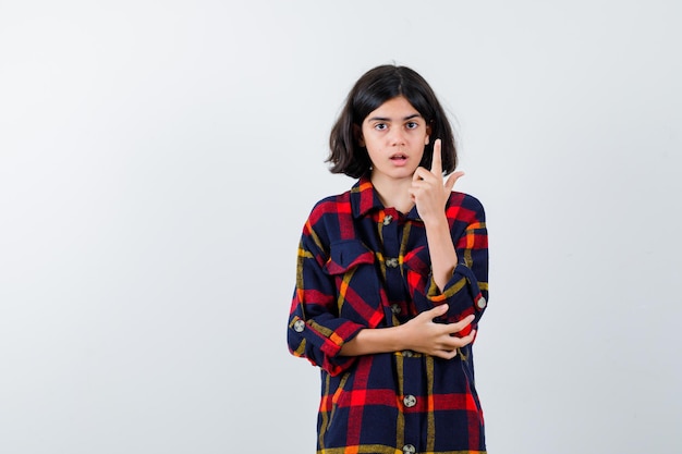 Chica joven en camisa a cuadros levantando el dedo índice en gesto de eureka mientras sostiene la mano en el codo y se ve sensible, vista frontal.