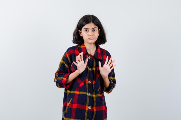Chica joven en camisa a cuadros levantando las manos en pose de rendición y mirando asustado, vista frontal.