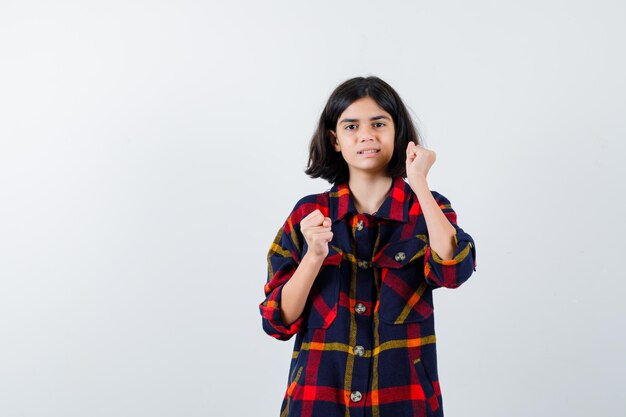 Chica joven en camisa a cuadros de pie en pose de boxeador y mirando poderosa, vista frontal.