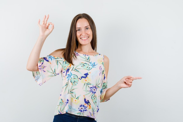 Chica joven en camisa, jeans apuntando hacia un lado con el signo de ok y mirando alegre, vista frontal.