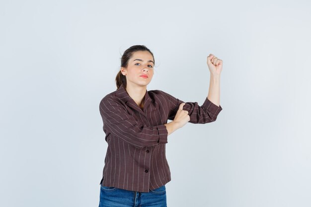 Chica joven en camisa a rayas, jeans mostrando los músculos del brazo, con la mano en el brazo y mirando serio, vista frontal.
