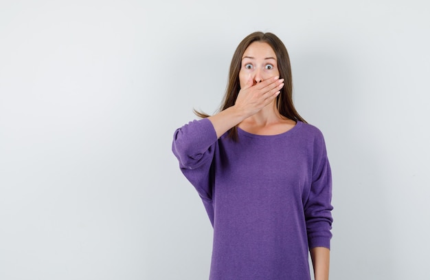 Chica joven en camisa violeta cubriendo la boca con la mano y mirando asustada, vista frontal.