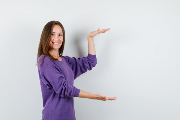 Chica joven en camisa violeta mostrando signo de tamaño y mirando alegre, vista frontal.