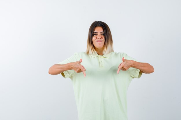 Chica joven en camiseta apuntando hacia abajo con los dedos índices y mirando linda, vista frontal.