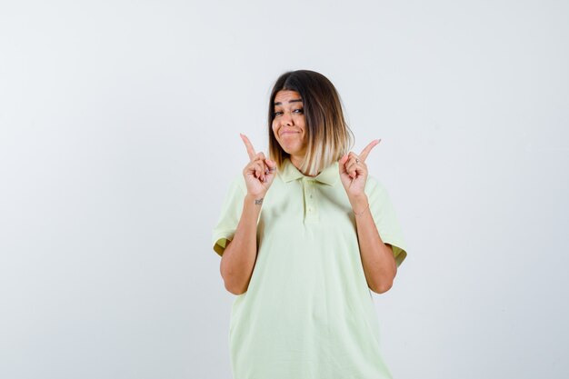 Chica joven en camiseta apuntando hacia arriba con los dedos índices y mirando feliz, vista frontal.