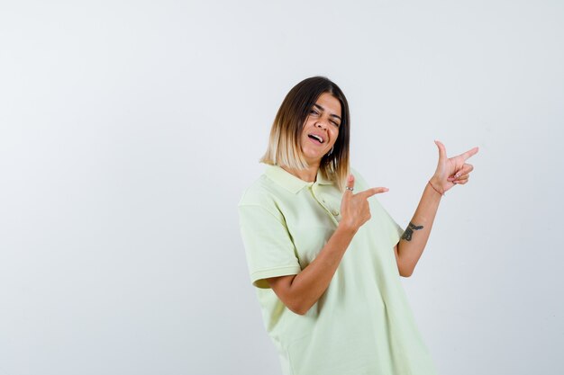 Chica joven en camiseta apuntando a la derecha con los dedos índices y mirando alegre, vista frontal.