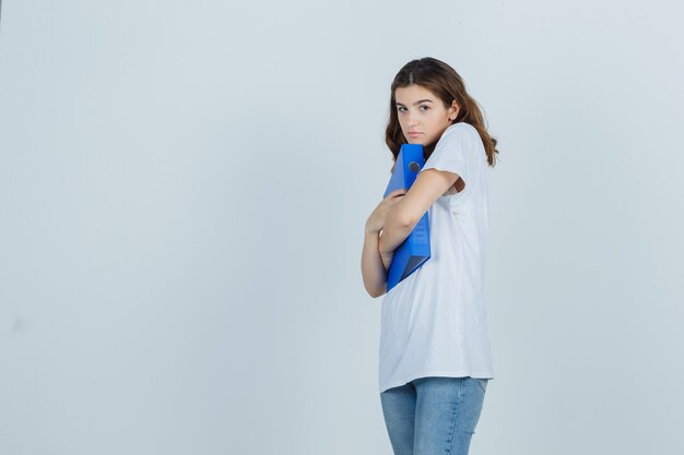 Chica joven en camiseta blanca abrazando la carpeta y mirando avergonzado, vista frontal.