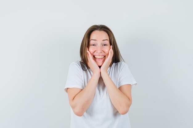 Chica joven en camiseta blanca acolchado la cara en sus manos y mirando alegre, vista frontal.