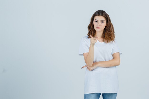 Chica joven en camiseta blanca apuntando a la cámara y mirando confiada, vista frontal.