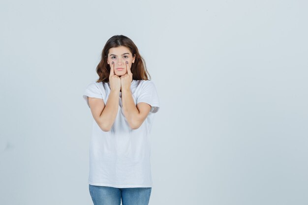 Chica joven en camiseta blanca apuntando a sus párpados y mirando sensible, vista frontal.