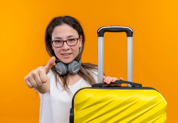 Chica joven en camiseta blanca con auriculares alrededor del cuello sosteniendo maleta de viaje sonriendo mostrando los pulgares para arriba